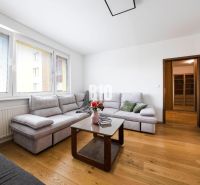 A bright living room with a corner sofa, wooden doors, and a floor with a wood decor.