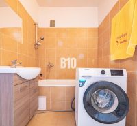A bathroom in a 3-room apartment with a washing machine, a bathtub, and light tiles.