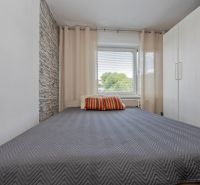 Bedroom of a 3-room apartment with a gray bed, brick cladding, and a large window.