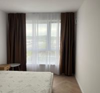 A bedroom in a 2-room apartment with a wood-patterned floor, simple furniture, and a large window.