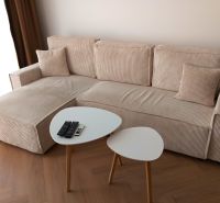 A light sofa with cushions, a pair of tables, a floor with a wooden decor, remote controls.