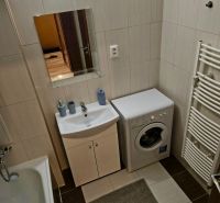 A bathroom in a studio apartment with a washing machine, sink, bathtub, and light-colored tiles.
