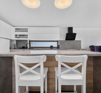 A kitchen in a family house with bar stools, white cabinets, and a wood-patterned floor.