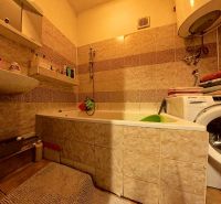 A bathroom in a family house with a bathtub, a washing machine, shelves, and light tiles.