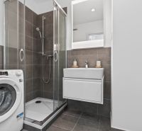 A bathroom in a studio apartment with a washing machine, shower, and sink with a lighted mirror.