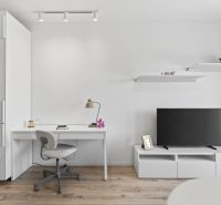 Studio apartment with white furniture, a desk, a television, and a floor with a wooden decor.