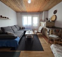 A living room of a cottage with a comfortable sofa, a tiled stove, and a wooden-patterned floor.