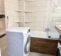 A bathroom in a 3-room apartment with a washing machine, bathtub, and shelving units.