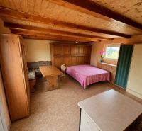 A room in a cottage with a wooden ceiling, a bed, a wardrobe, and a table with chairs.