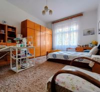 A room in a 4-room apartment with furniture, carpeted floor, bed, and a window with white curtains.