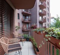 A balcony with chairs and flower pots in a 2-room apartment, overlooking a green area.