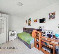 A room in a 3-room apartment with a single bed, a washing machine, and shelves. Floor with wood decor.