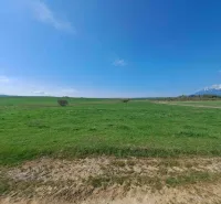 Green agricultural and forest lands near Podhorany with mountains in the distance under a blue sky.