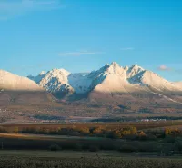 Poprad agricultural and forest lands with a panorama of snow-covered Tatra peaks under a blue sky.
