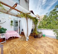 The terrace of a 2-room apartment with a wooden decor floor, sheltered by greenery and decorative lamps.