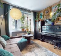 Living room of a two-room apartment with a piano, sofa, houseplants, and a wooden-patterned floor.