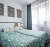 A bedroom with a large bed and geometric paintings, curtains letting in daylight.