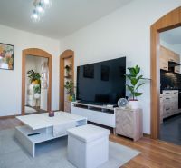 Living room of a two-room apartment with a large television, yellow sofa, and wooden decor flooring.