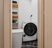 Laundry room in a 3-bedroom apartment with a washing machine and a wooden shelf, complemented by a dark floor.