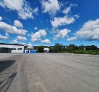 Warehouses and halls in Ličartovce with a paved area and blue containers under a blue sky.
