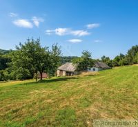 The cabin in Dobroč, surrounded by trees, lies on a green slope under a blue sky.