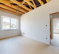 The interior of a family house with bare walls, a large window, and exposed ceiling beams.