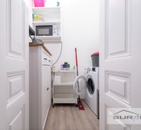 Laundry room in a 2-room apartment with a washing machine, shelf, microwave, and wooden decor flooring.