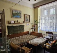 Living room of a two-room apartment with a sofa, paintings, and a large window with curtains.