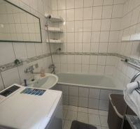 A bathroom in a 2-room apartment with a bathtub, sink, and washing machine, tiled with white tiles.