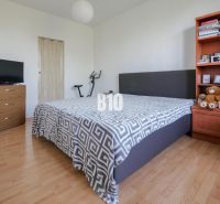 Bedroom in a two-room apartment with a wood-patterned floor, bed, and shelf.