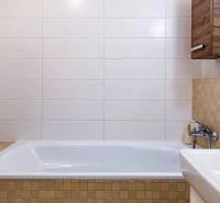 Bathroom in a 2-room apartment with white tiles and a bathtub, wooden decor on the cabinet.