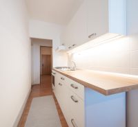 A narrow kitchen with white cabinets, tiled backsplash, and wood-patterned flooring in a two-room apartment.