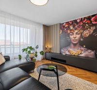 Living room with a leather sofa, artistic wallpaper, and wood-patterned flooring in a three-room apartment.