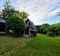 A cottage in Podhorany surrounded by greenery, with a blue sky and clouds in the background.