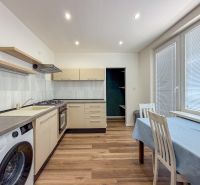 A kitchen in a 2-room apartment with appliances, a dining table, and a wooden decor floor.