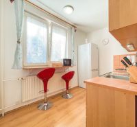 The kitchen of a one-room apartment with a wooden decor floor, red chairs, and a window.