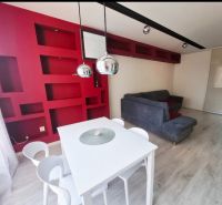 The interior of a 3-room apartment with red shelves, a white table, and a gray sofa.