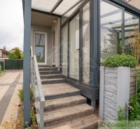 A cottage in Senec with a glazed veranda, staircase, and paved courtyard, bordered by greenery.