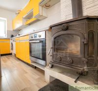 A kitchen in a cottage with an orange kitchen unit and a tiled stove, and a floor with a wooden decor.