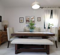 Dining room with a wooden table, upholstered chairs, decorative paintings, light curtains, wooden sideboard.