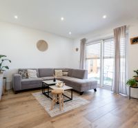 Living room in a family house with a gray sofa, wood-patterned flooring, and large windows.