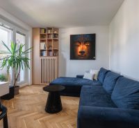 Living room with a blue sofa, painting, bookshelf, and wooden decor flooring in a three-room apartment.