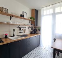 A kitchen in a 3-room apartment with black cabinets, wooden shelves, and a tiled floor.