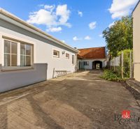 A family house in Budmerice with a paved courtyard, next to a fence with vegetation.
