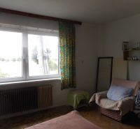 Interior of a family house with an armchair, bed, shelf, and large window.