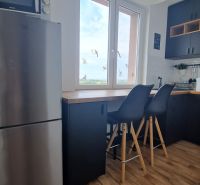 The kitchen of a 2-room apartment with a wooden decor floor, bar stools, and a window.