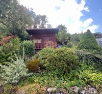 A wooden cabin in the green gardens of Ružomberok surrounded by trees and shrubs under a clear sky.
