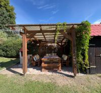 A wooden pergola in front of the cottage in Beňadiková surrounded by greenery and a sunny yard.