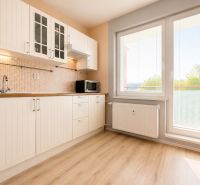 A kitchen in a one-room apartment with a wooden decor floor, large windows, and white cabinets.