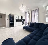 Living room with a dark blue couch, kitchen, and wooden decor flooring in a 3-room apartment.